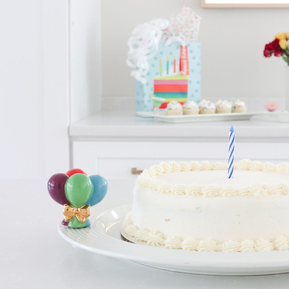A ceramic mini four colorful balloons wrapped in a gold bow on a round platter that holds a white birthday cake with one candle.