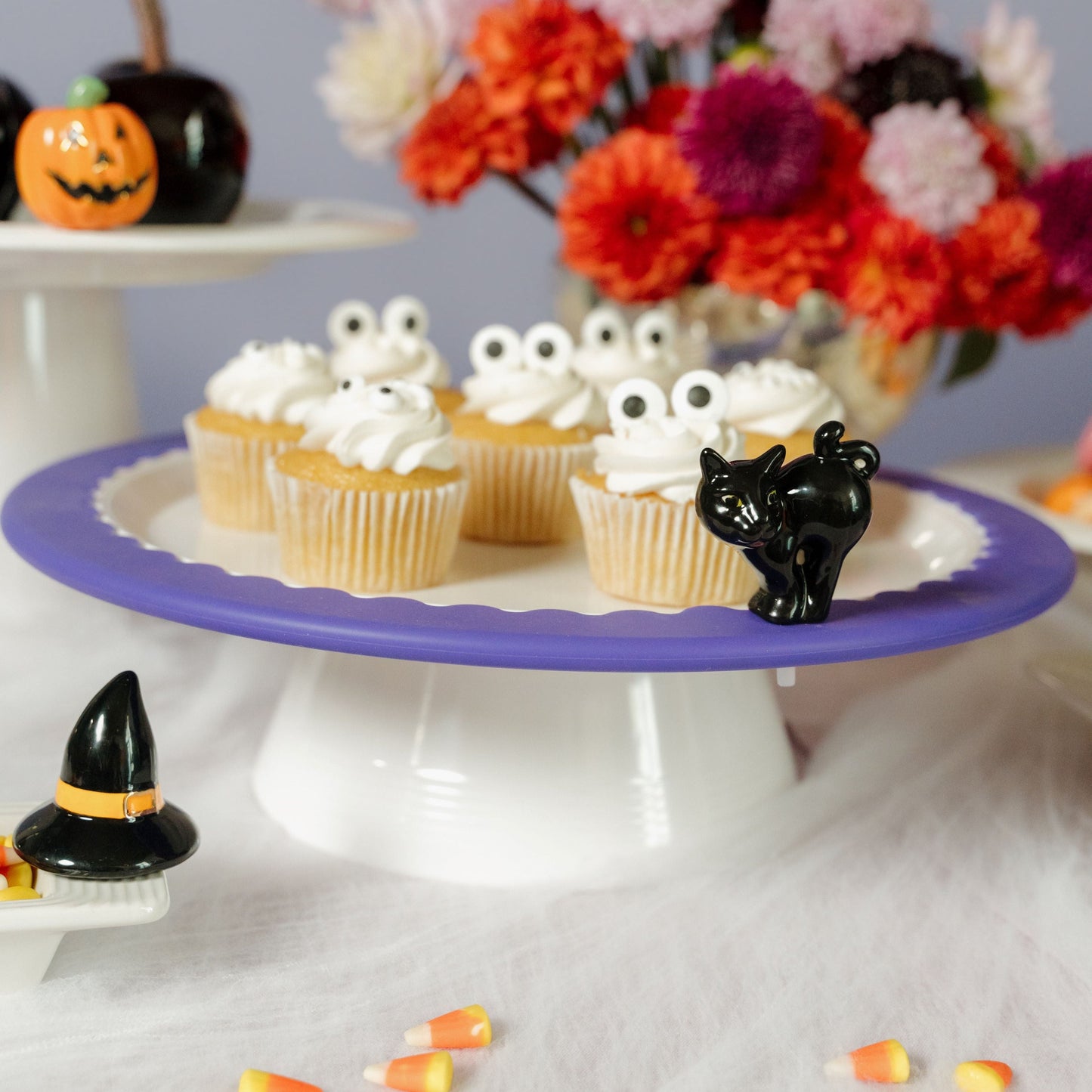 A white round melamine riser with a central screw hole, designed to hold a platter. The round platter is holding ghost cupcakes. There is a black cat mini on top of the round platter.
