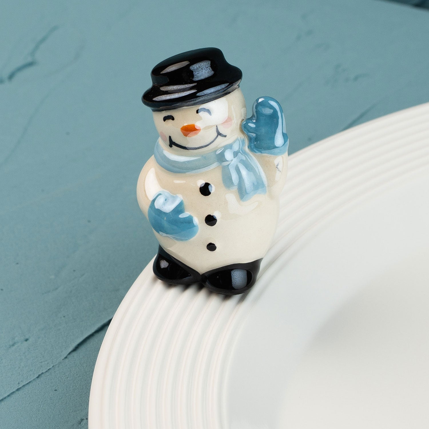 snowman mini with a black top hat, blue scarf, and gloves on a white plate against a teal background.